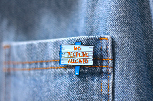 A close-up of a small enamel pin shaped like a wooden sign that reads 'NO PEOPLING ALLOWED' in orange lettering, clipped onto the pocket of blue denim jeans.
