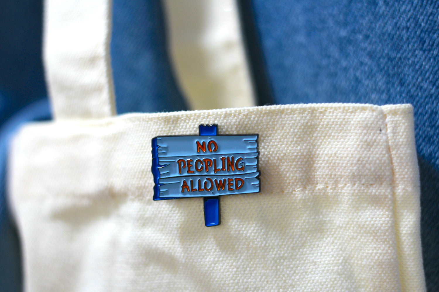 A close-up of a canvas tote bag with a blue enamel pin shaped like a street sign that reads 'NO PECCING ALLOWED' in red lettering, set against a blue denim background.