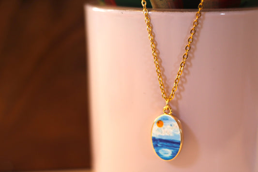 A gold chain necklace with an oval pendant featuring a painted seascape scene, hanging against a pale pink cylindrical background.
