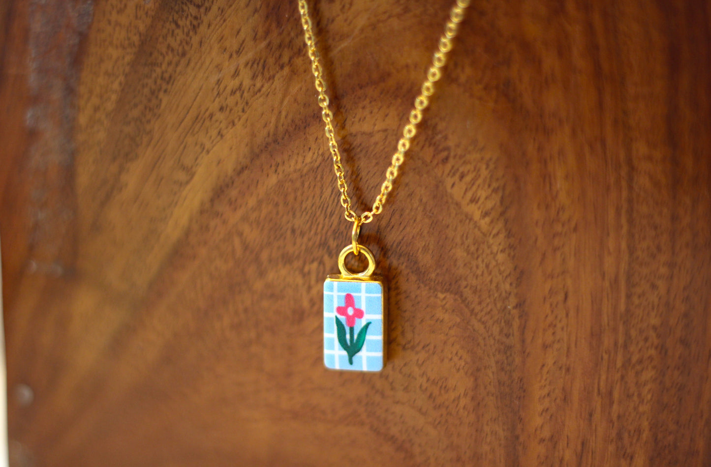 A gold chain necklace with a small rectangular pendant featuring a light blue and white checkered pattern, a pink flower, and green leaves, displayed on a wooden surface.