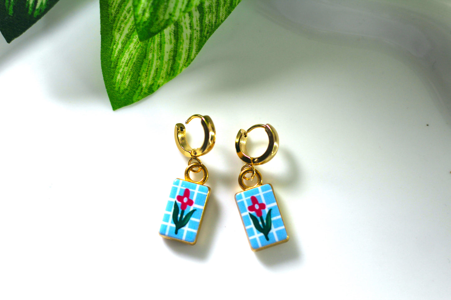 A pair of gold hoop earrings with rectangular blue-and-white checkered dangles featuring a small red flower and green stem, photographed on a white surface with a green leaf in the background.