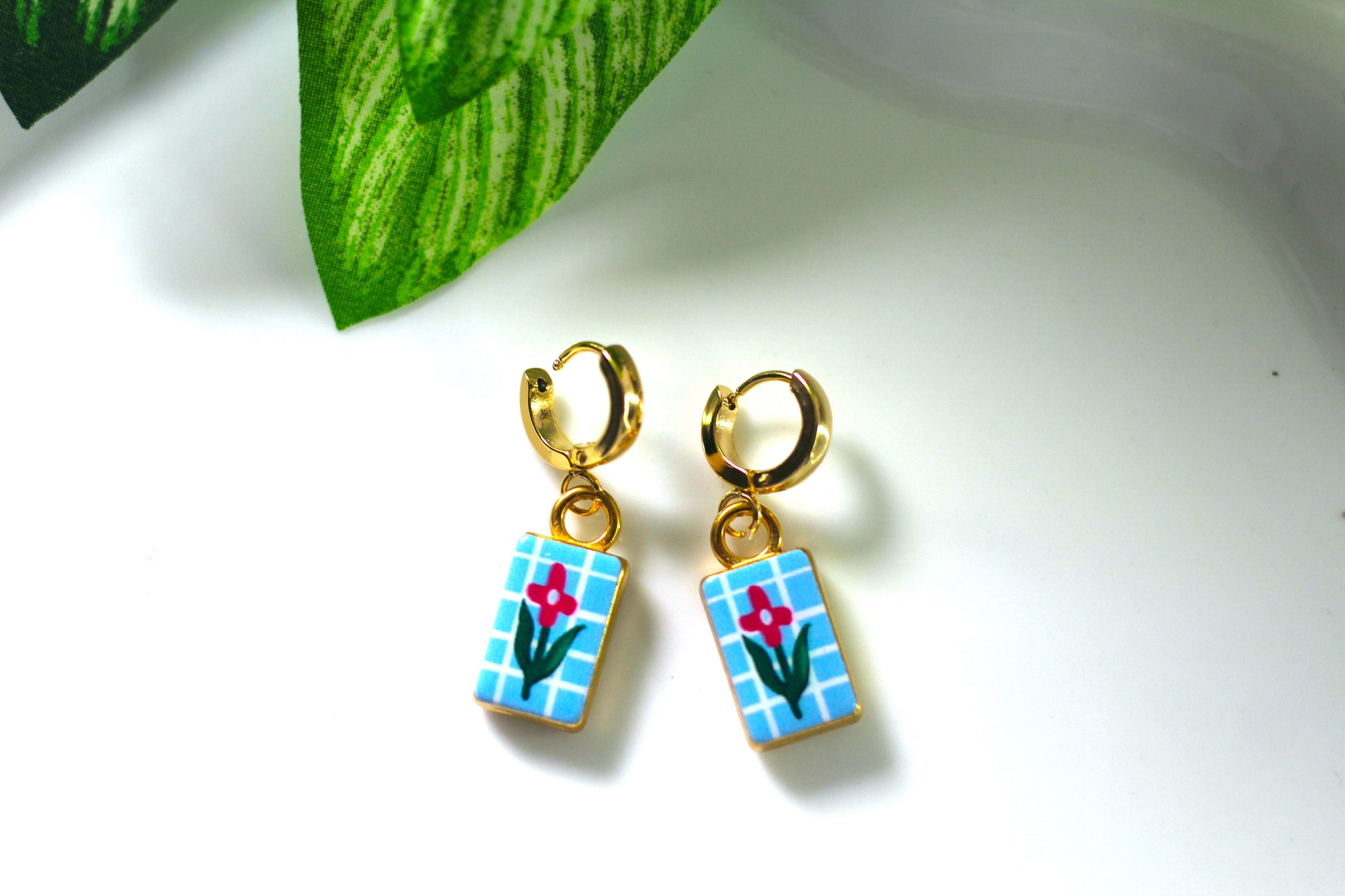 A pair of gold hoop earrings with rectangular blue-and-white checkered dangles featuring a small red flower and green stem, photographed on a white surface with a green leaf in the background.