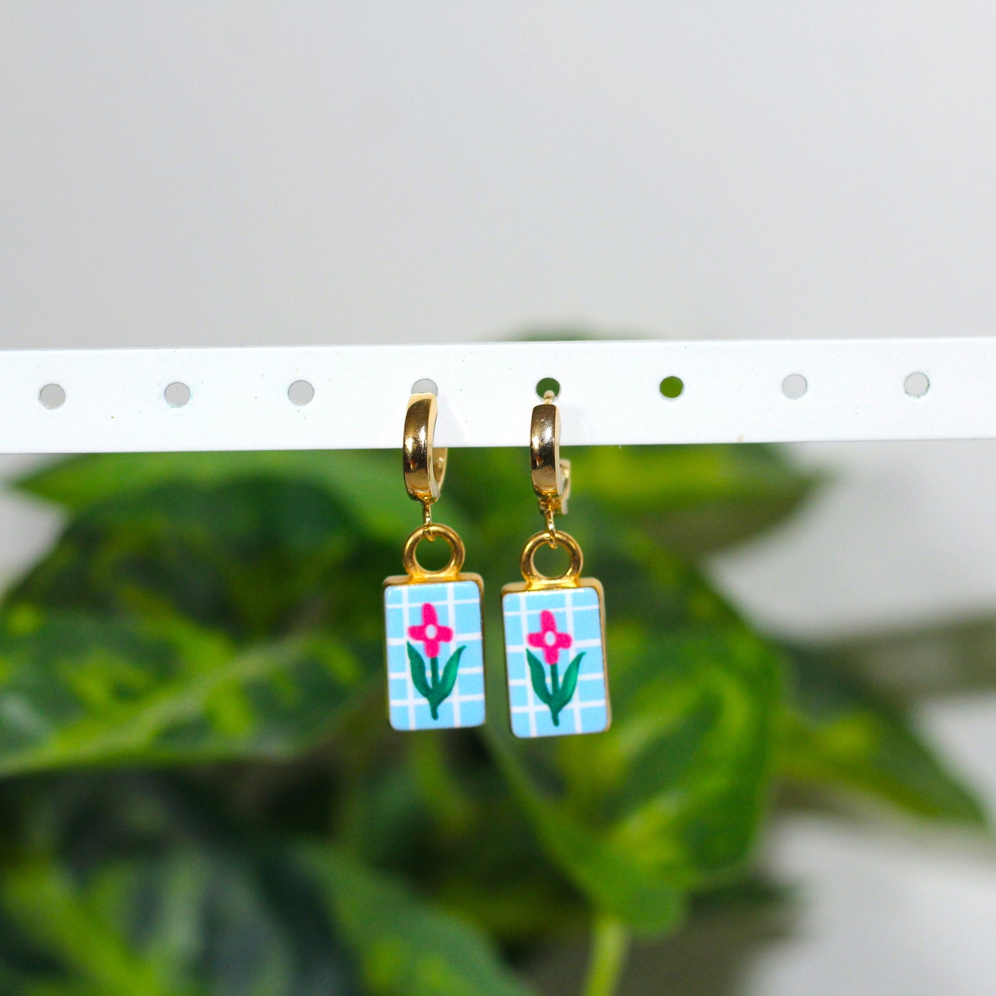A pair of gold hoop earrings with rectangular blue-and-white plaid dangles featuring pink flowers and green stems, hanging from a white display bar with blurred green leaves in the background.