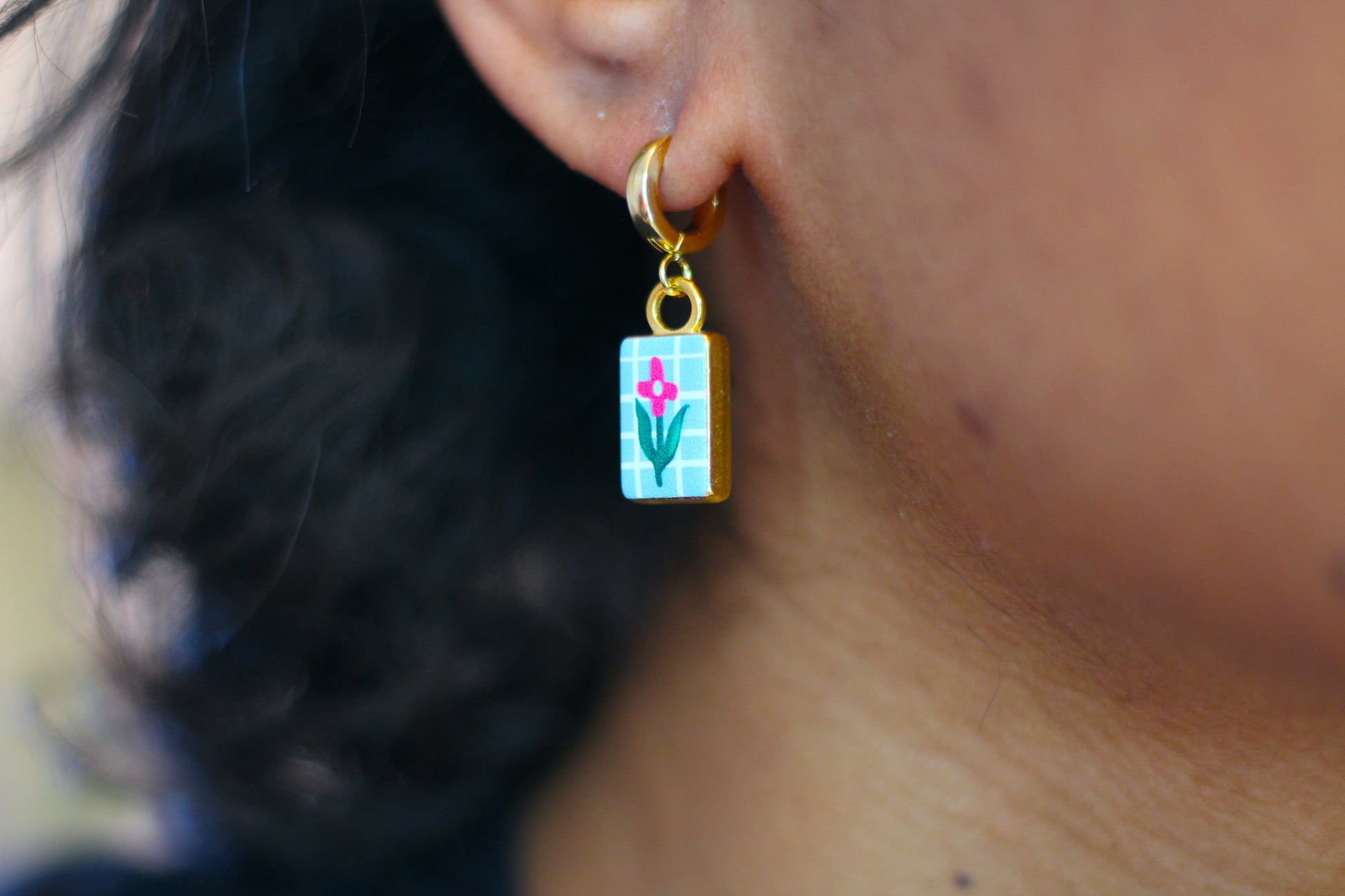 A close-up of a person's ear wearing a small gold hoop earring with a rectangular charm featuring a pink flower on a light blue grid background.