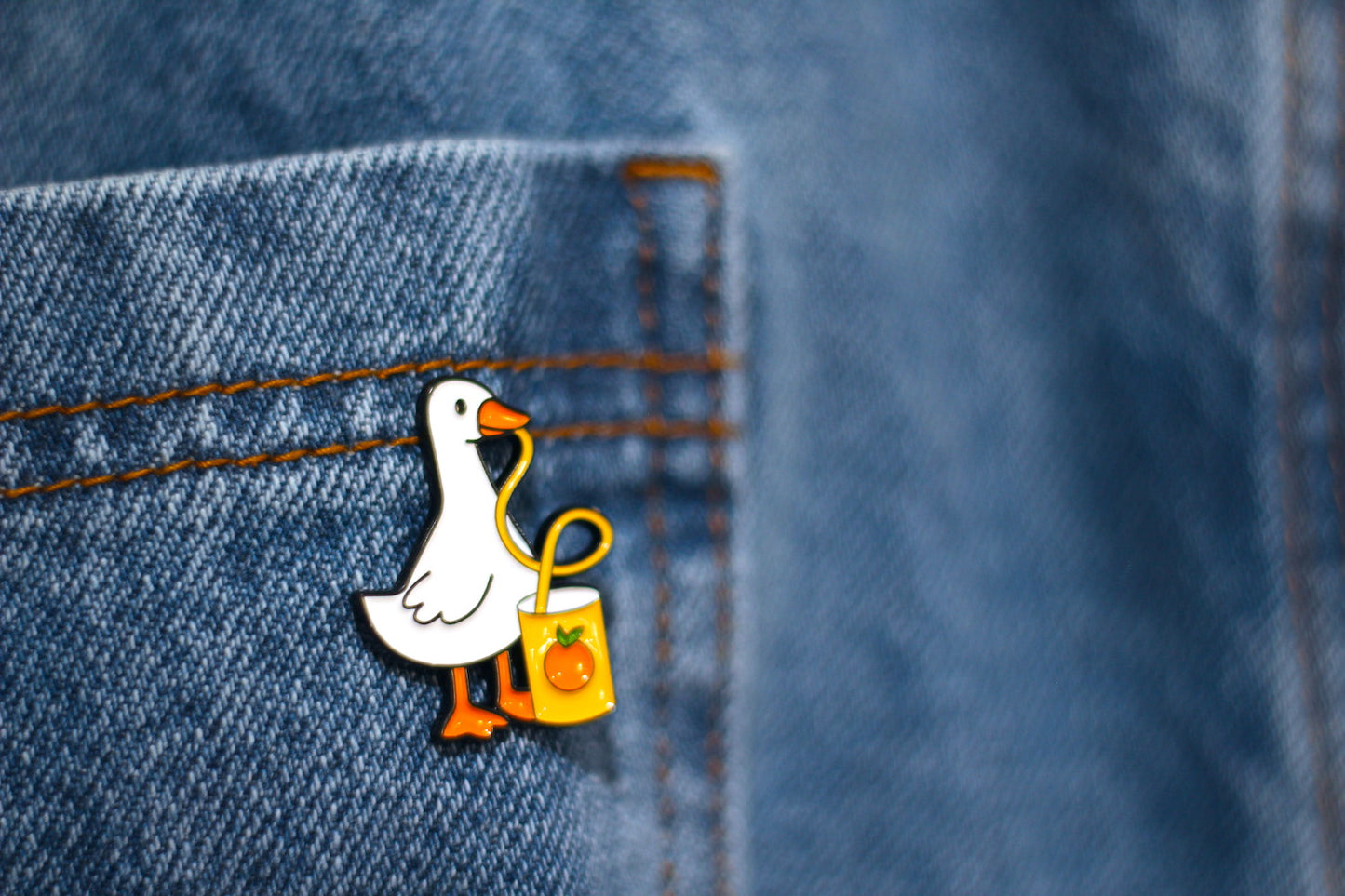 A close-up of a white duck enamel pin with orange beak and feet, holding a yellow bag, pinned onto blue denim fabric.