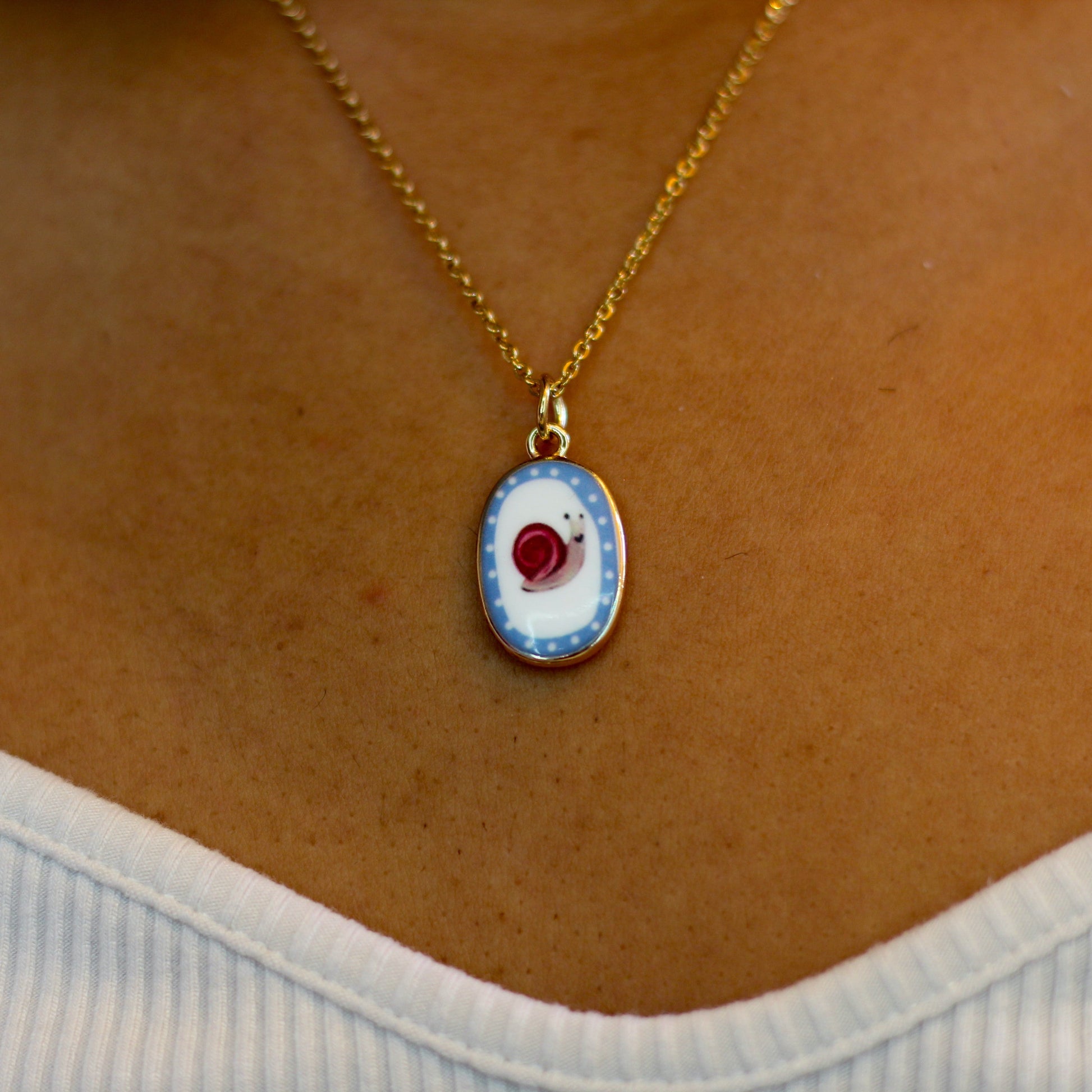A gold chain necklace with an oval pendant featuring a blue and white pattern and a snail, worn on a person's neck over a white ribbed top.