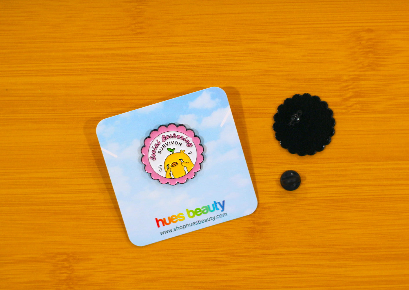 A small round enamel pin with a pink flower border and a yellow cartoon duck, mounted on a light blue backing card that says "social gathering survivor". The backing card is placed on a warm wood surface, with a back view of the pin visible to the right.