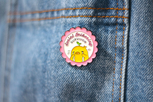A circular enamel pin featuring a yellow cartoon suck and the text 'Social Gathering SURVIVOR' on a pink scalloped border, pinned onto blue denim fabric.