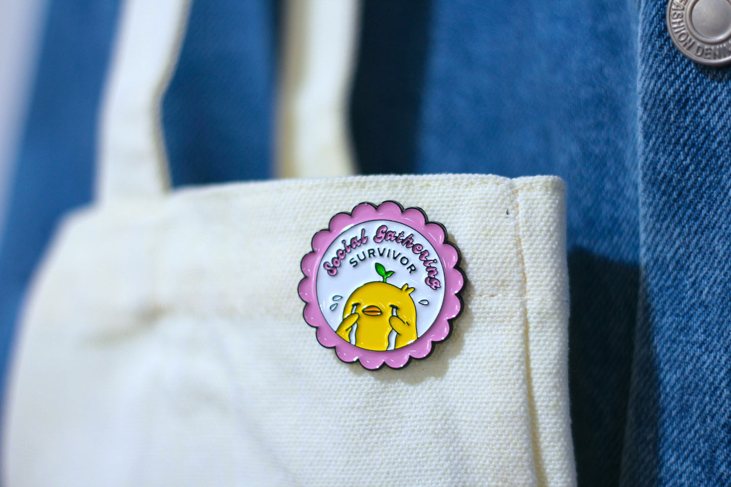 A circular enamel pin featuring a yellow crying character with the text 'Social Gathering SURVIVOR' on a pink scalloped border, pinned onto a cream canvas tote bag next to a blue denim jacket.