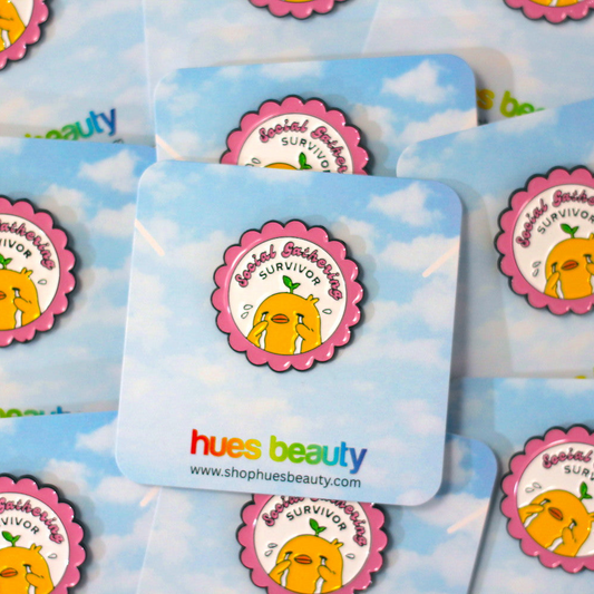 A close-up of multiple square cards featuring the same circular pin design with a crying duck that says 'Social Gathering Survivor". The cards have a light blue sky-and-cloud print and the brand name 'hues beauty' with the website printed at the bottom.