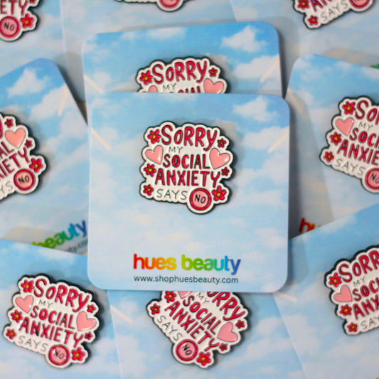 Several blue sky-themed cards with pink and white 'Sorry My Social Anxiety Says No' enamel pins are arranged on a light surface. The center card shows the brand name 'hues beauty'.