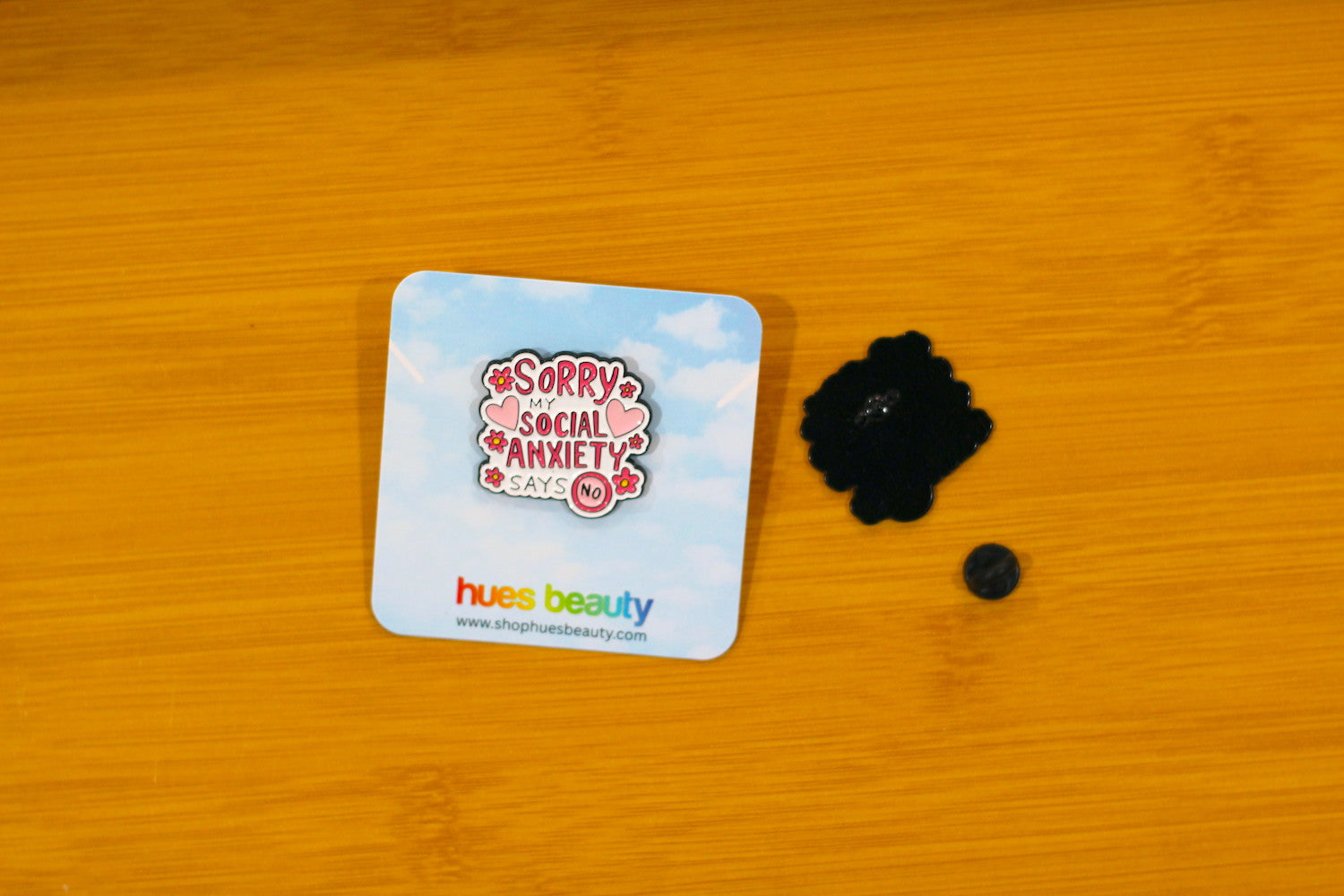 A small black flower-shaped pin with a central button and a tiny matching black bead, shown next to its blue backing card on a wooden surface. The enamel pin features the text 'Sorry Social Anxiety' and the brand card says 'hues beauty'.