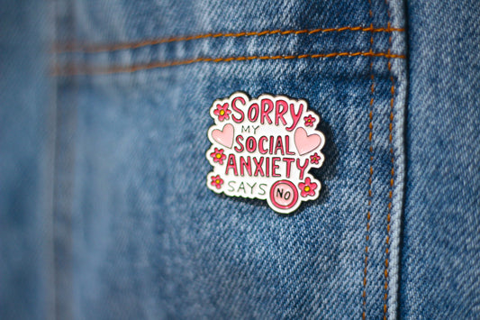 A close-up of a enamel pin attached to blue denim jeans. The pin reads 'Sorry My Social Anxiety Says No' in pink and white lettering with pink hearts and flowers.