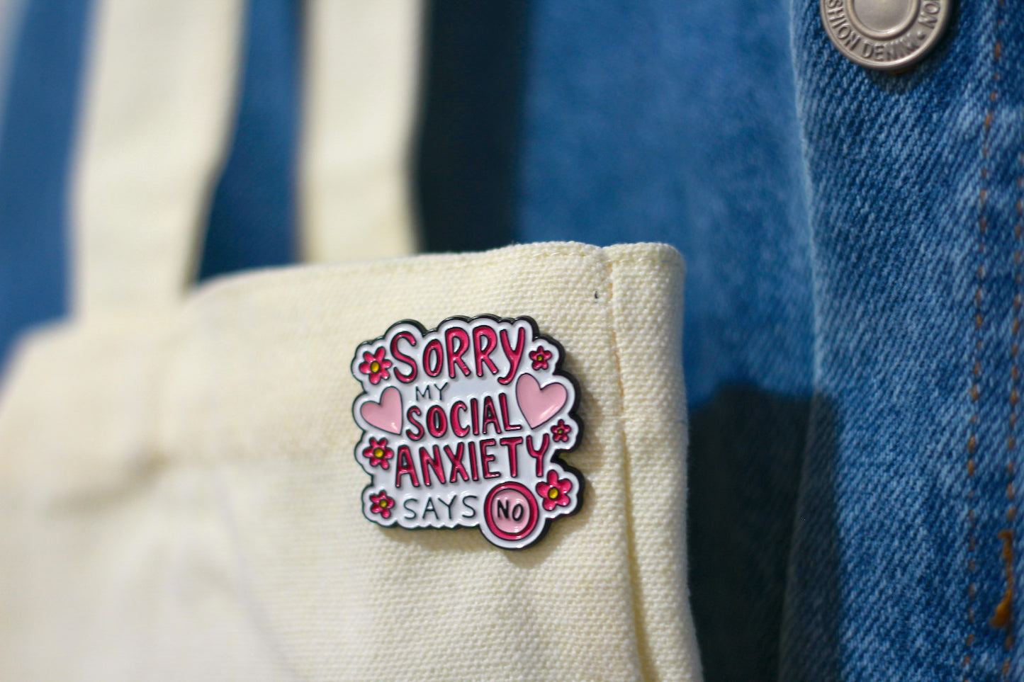 A close-up of a small enamel pin attached to a cream canvas tote bag, with a blue denim jacket in the background.