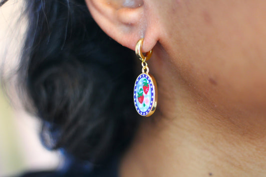 A close-up of a person's ear wearing a gold huggie hoop earring with a colorful oval charm featuring a strawberry design.