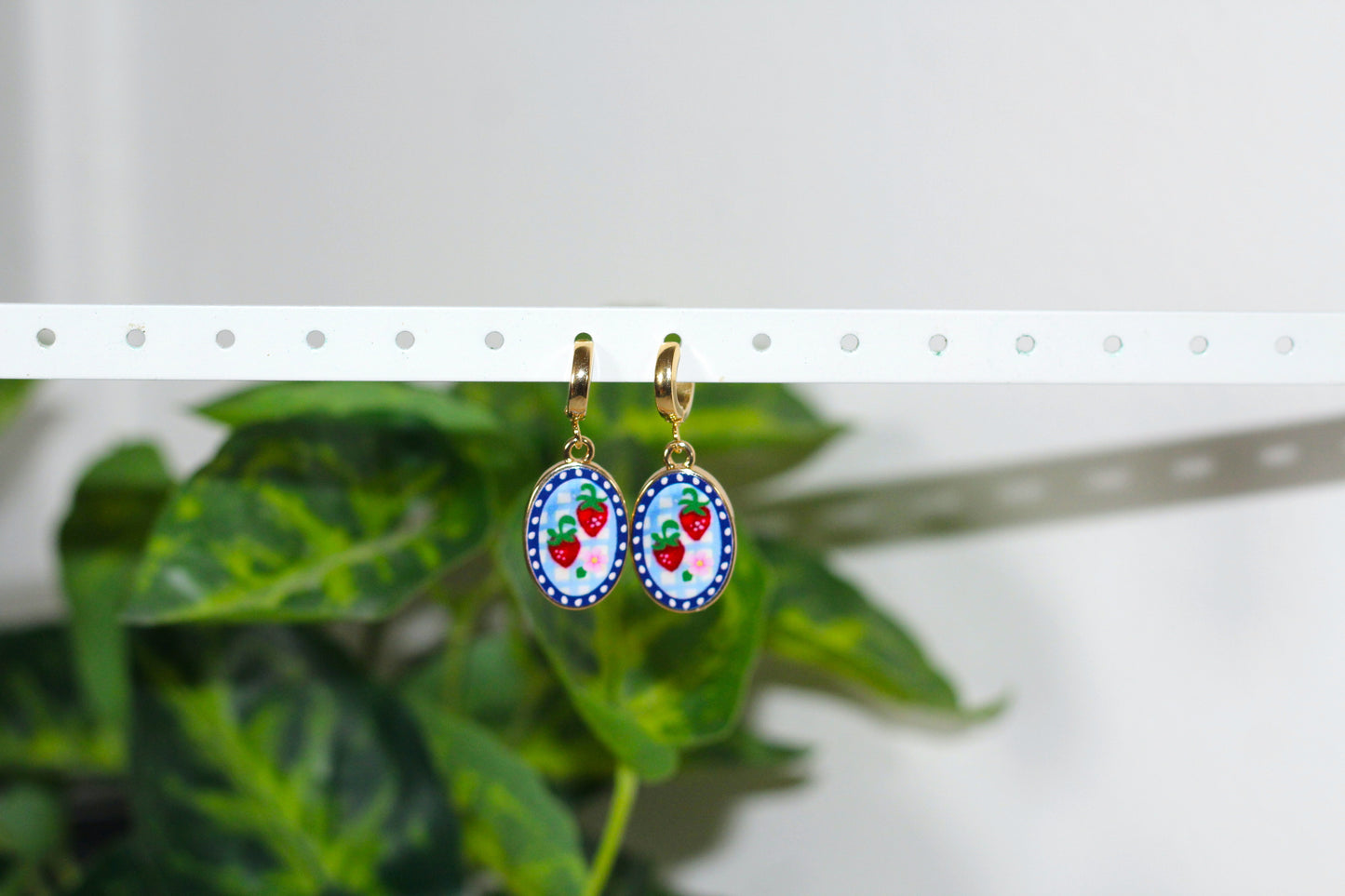 A pair of oval dangling earrings with colorful strawberry-themed designs, hanging from a white peg display bar in front of blurred green leaves.