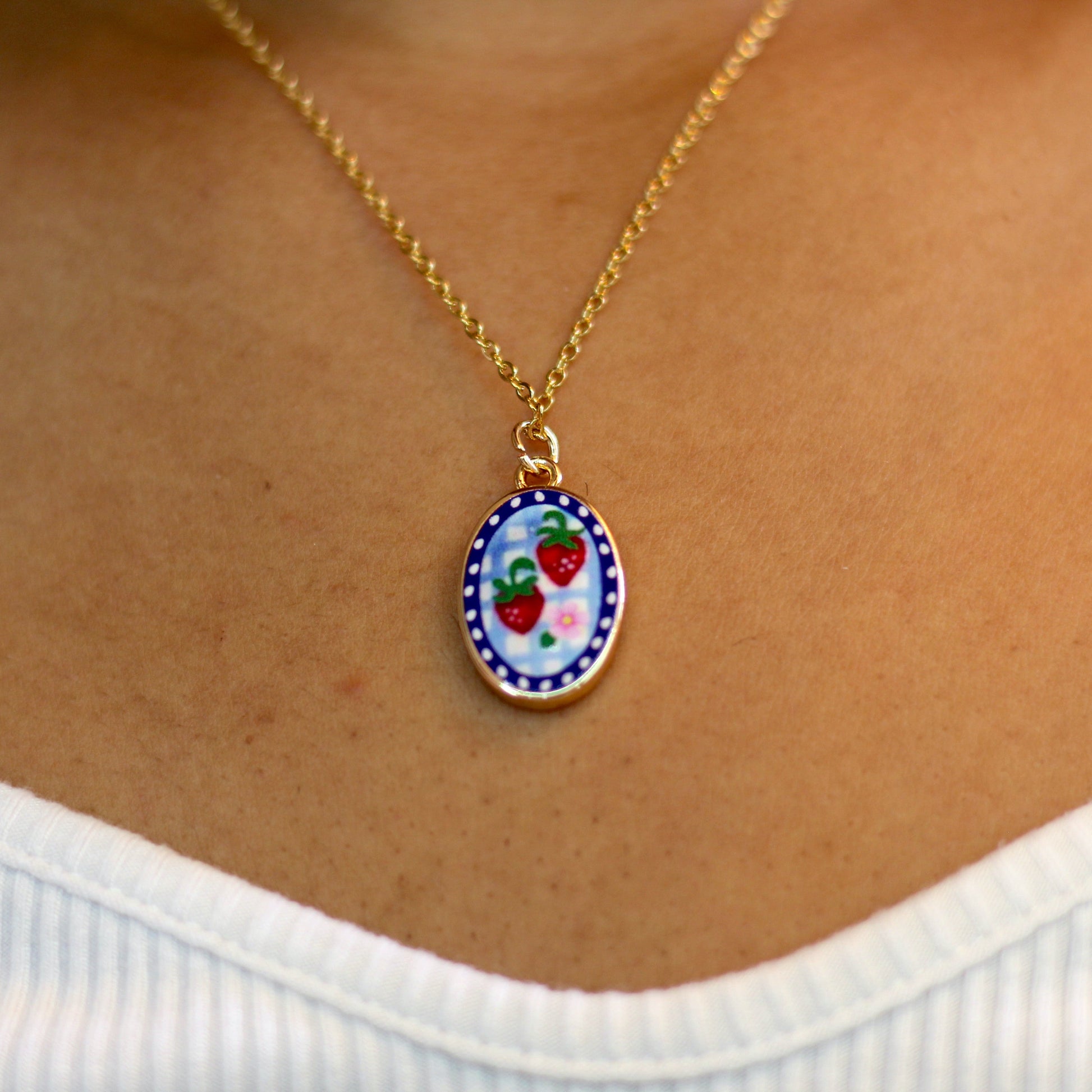 A gold chain necklace with an oval pendant featuring a colorful strawberry design, worn on a person's neck over a white ribbed top.