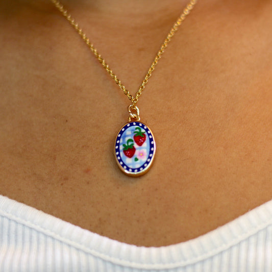 A gold chain necklace with an oval pendant featuring a colorful strawberry design, worn on a person's neck over a white ribbed top.