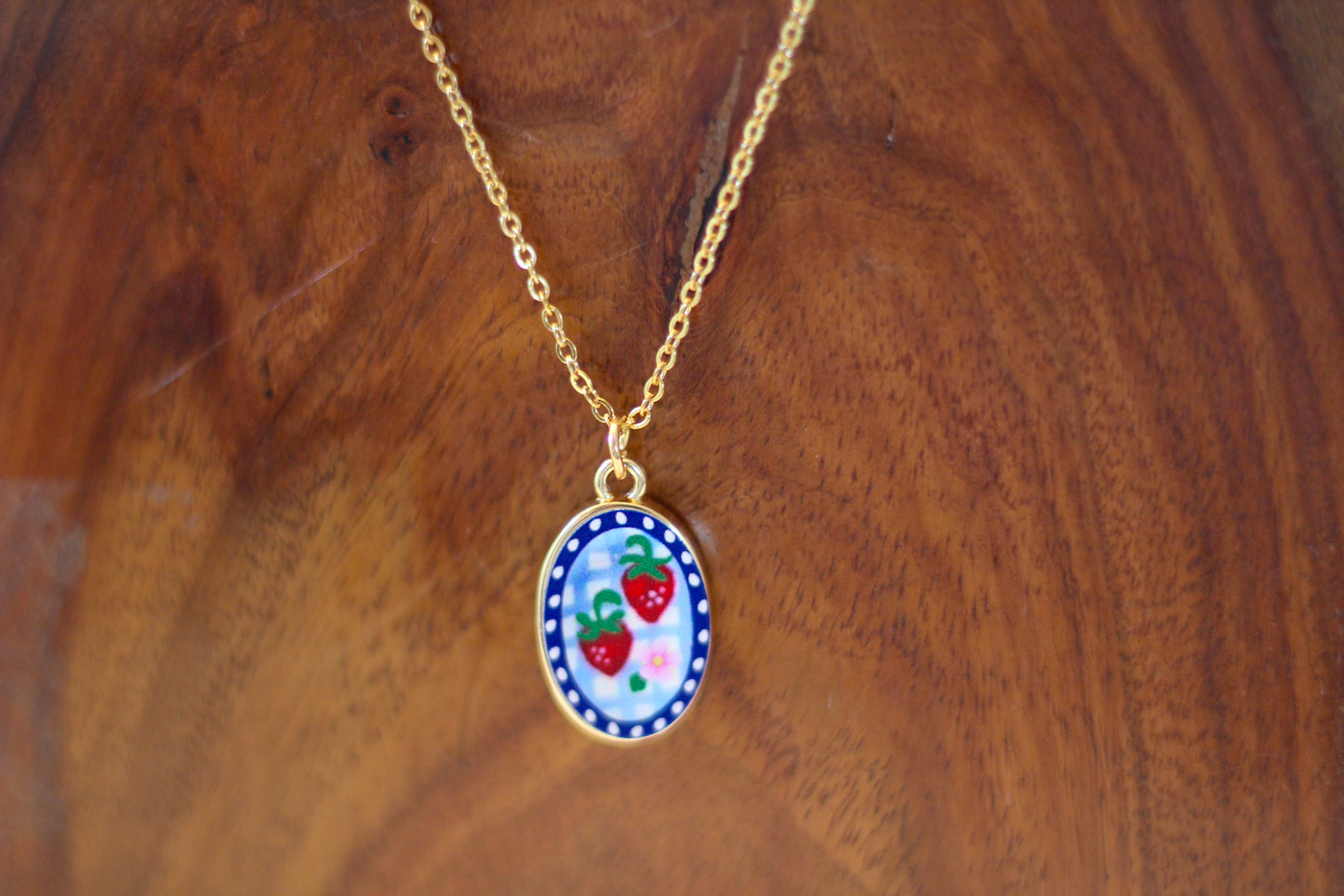 A gold chain necklace with an oval pendant featuring a white background decorated with red strawberries, green leaves, and a blue patterned border, displayed on a wooden surface.