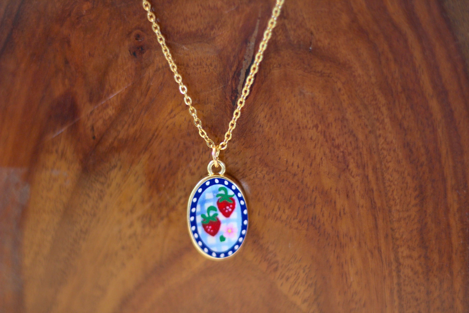 A gold chain necklace with an oval pendant featuring a white background decorated with red strawberries, green leaves, and a blue patterned border, displayed on a wooden surface.
