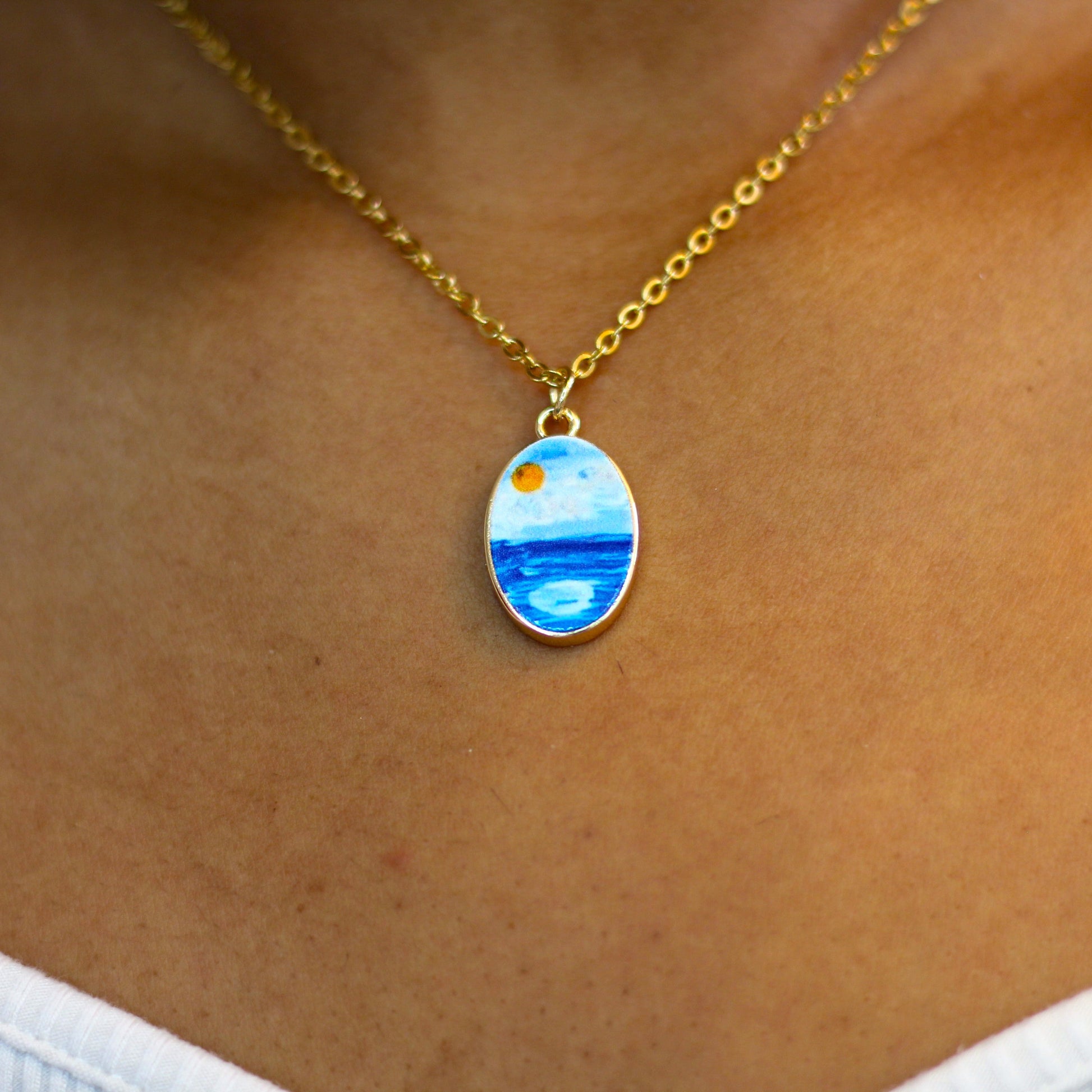 A gold chain necklace with an oval pendant featuring a painted landscape of blue water and a yellow sun, shown worn on a person's neck.