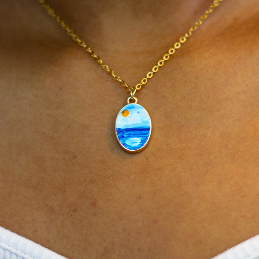 A gold chain necklace with an oval pendant featuring a painted landscape of blue water and a yellow sun, shown worn on a person's neck.