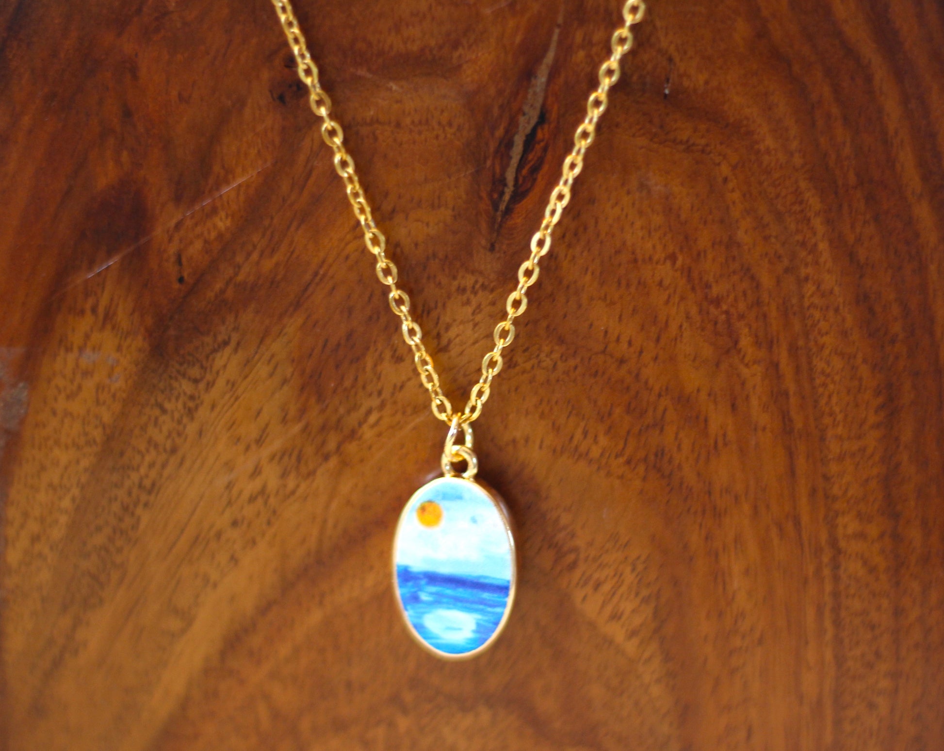 A gold chain necklace with an oval pendant featuring a painted blue and white landscape with a small yellow sun, displayed on a wooden surface.