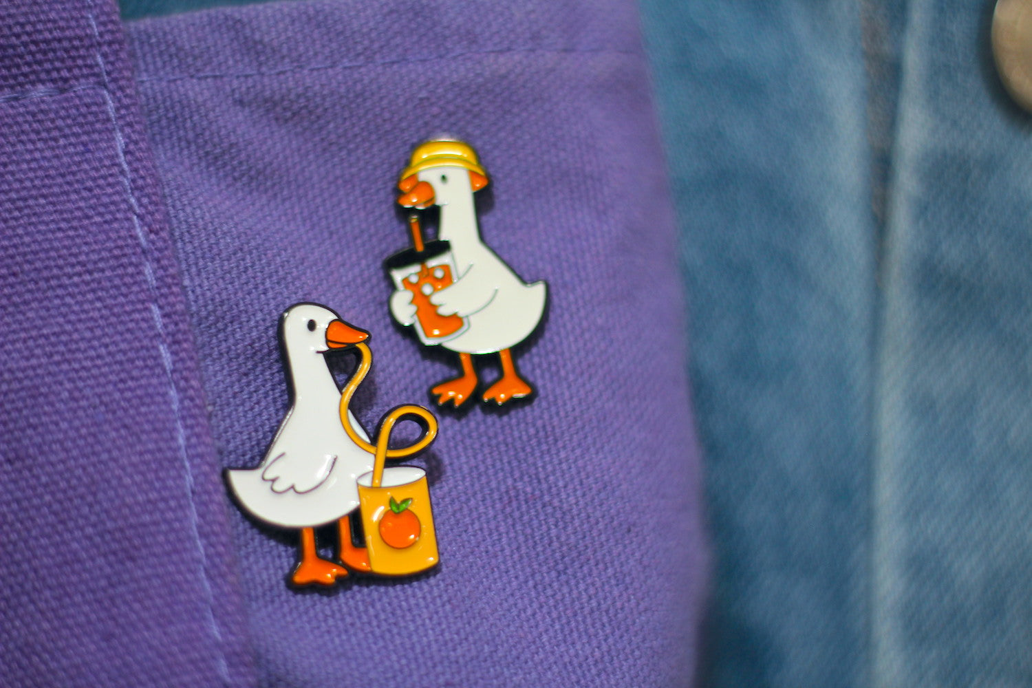 Two small duck enamel pins displayed on a purple fabric pocket next to a blue denim jacket.